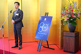 180205OSG賀詞交歓会3 閉会のあいさつをする大沢常務と80周年記念ロゴ。